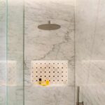Detalle de ducha de mármol blanco veteado con rociador metálico, hornacina de azulejos y mampara de cristal