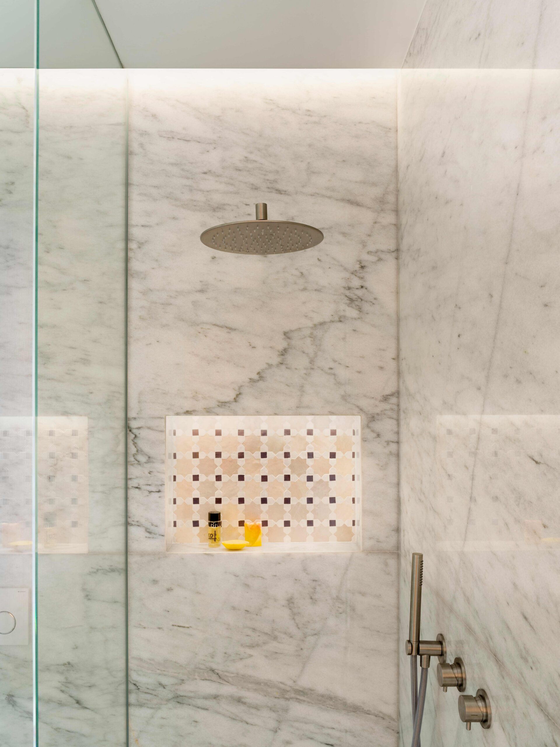 Detalle de ducha de mármol blanco veteado con rociador metálico, hornacina de azulejos y mampara de cristal