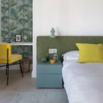 Dormitorio con cabecero verde tapizado, papel pintado de palmeras, escritorio empotrado y silla amarilla