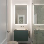Baño con mueble suspendido verde, espejo retroiluminado, ducha de cristal y puf de terciopelo verde