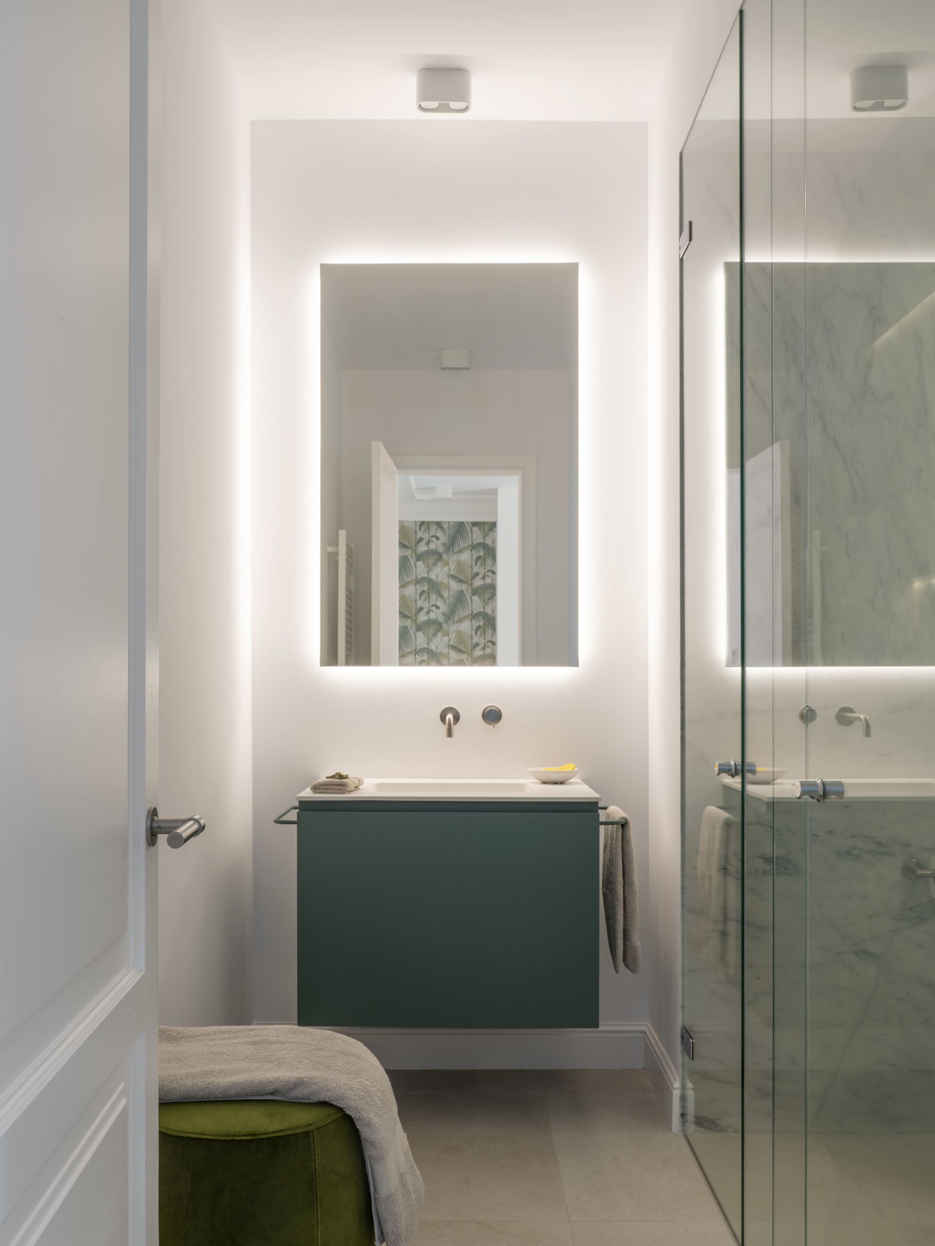 Baño con mueble suspendido verde, espejo retroiluminado, ducha de cristal y puf de terciopelo verde