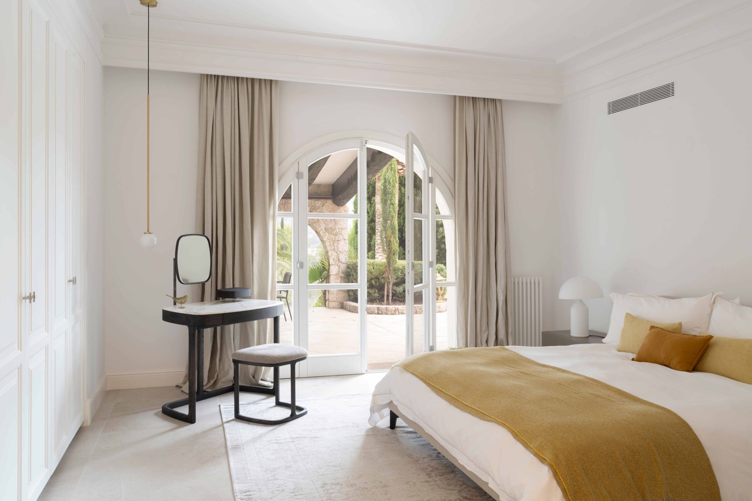 Dormitorio con puertas balconeras en arco al patio, tocador negro, cortinas beige y manta mostaza
