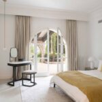 Schlafzimmer mit Bogentüren zum Garten, Vorhängen in Beige, schwarzem Schminktisch, senfgelber Decke und weißen Schränken
