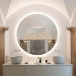 Baño con dos lavabos de mármol sobre mueble suspendido y espejo redondo retroiluminado bajo bóveda blanca