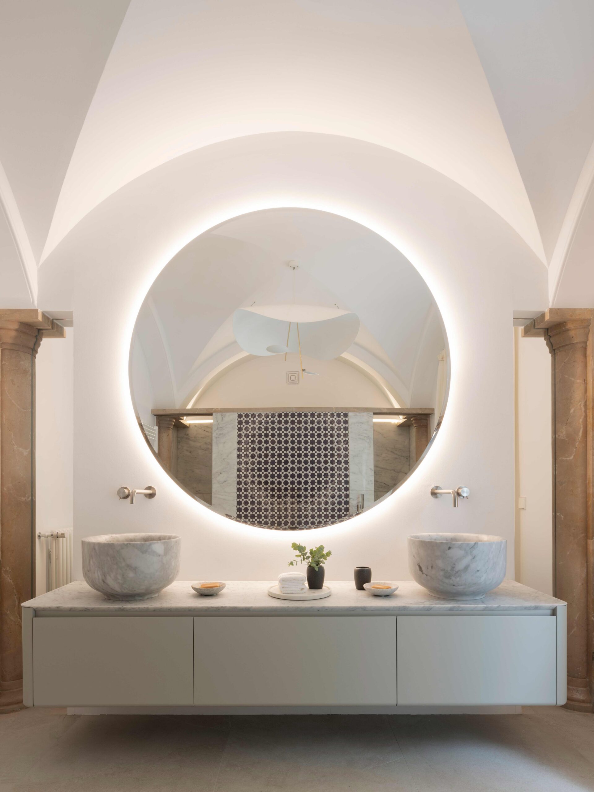 Baño con dos lavabos de mármol sobre mueble suspendido y espejo redondo retroiluminado bajo bóveda blanca