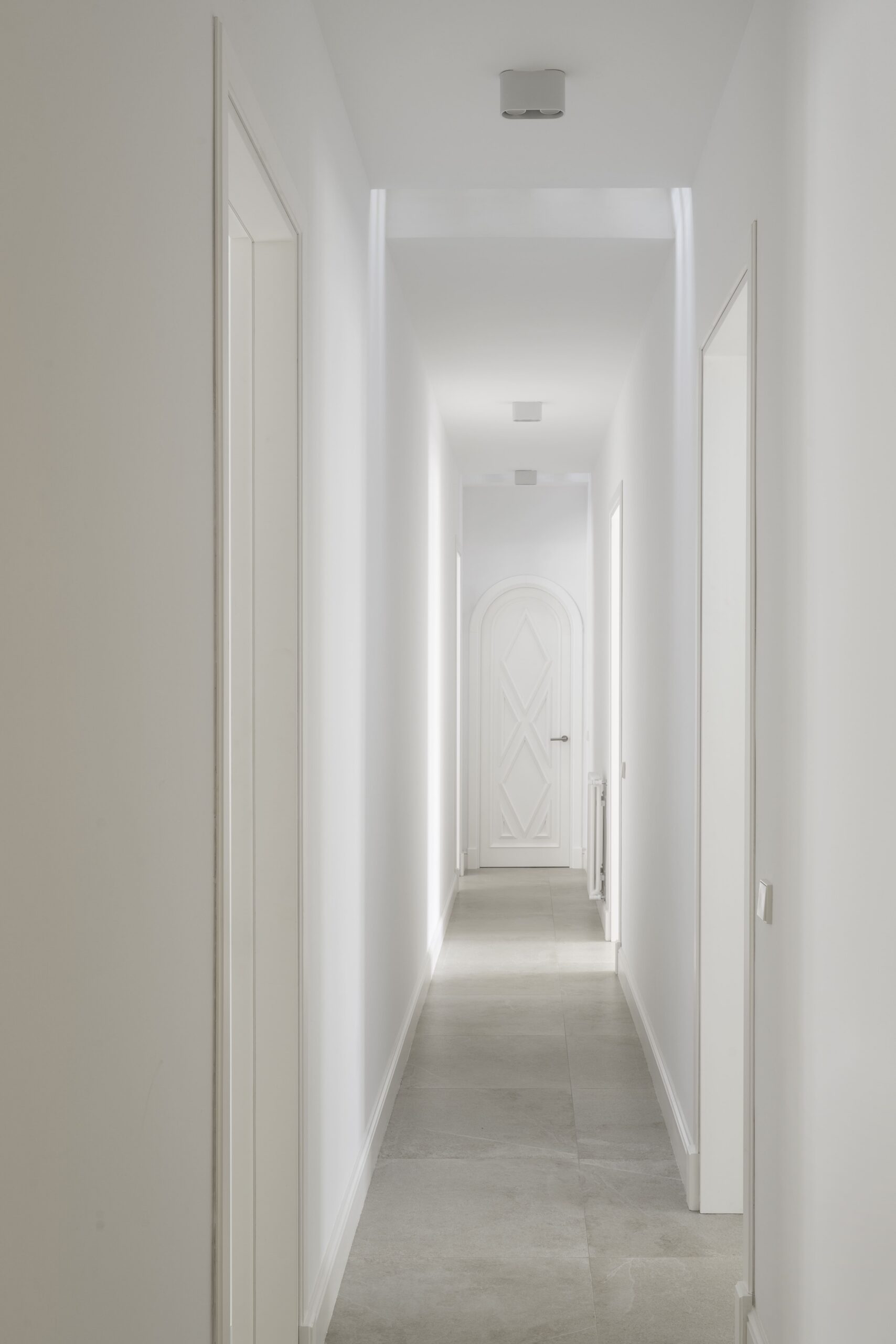 Pasillo blanco con suelo gris y puerta en arco con relieve de rombos al fondo