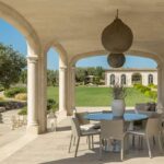 Veranda mit Steinbögen, rundem Tisch mit Stühlen, kugelförmigen Hängelampen und Blick in den Garten