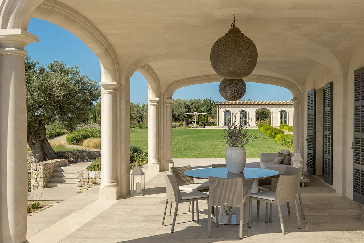 Veranda mit Steinbögen, rundem Tisch mit Stühlen, kugelförmigen Hängelampen und Blick in den Garten