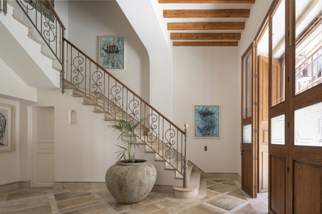 Vestíbulo con escalera de peldaños de piedra, barandilla de forja, gran tinaja y puertas de madera