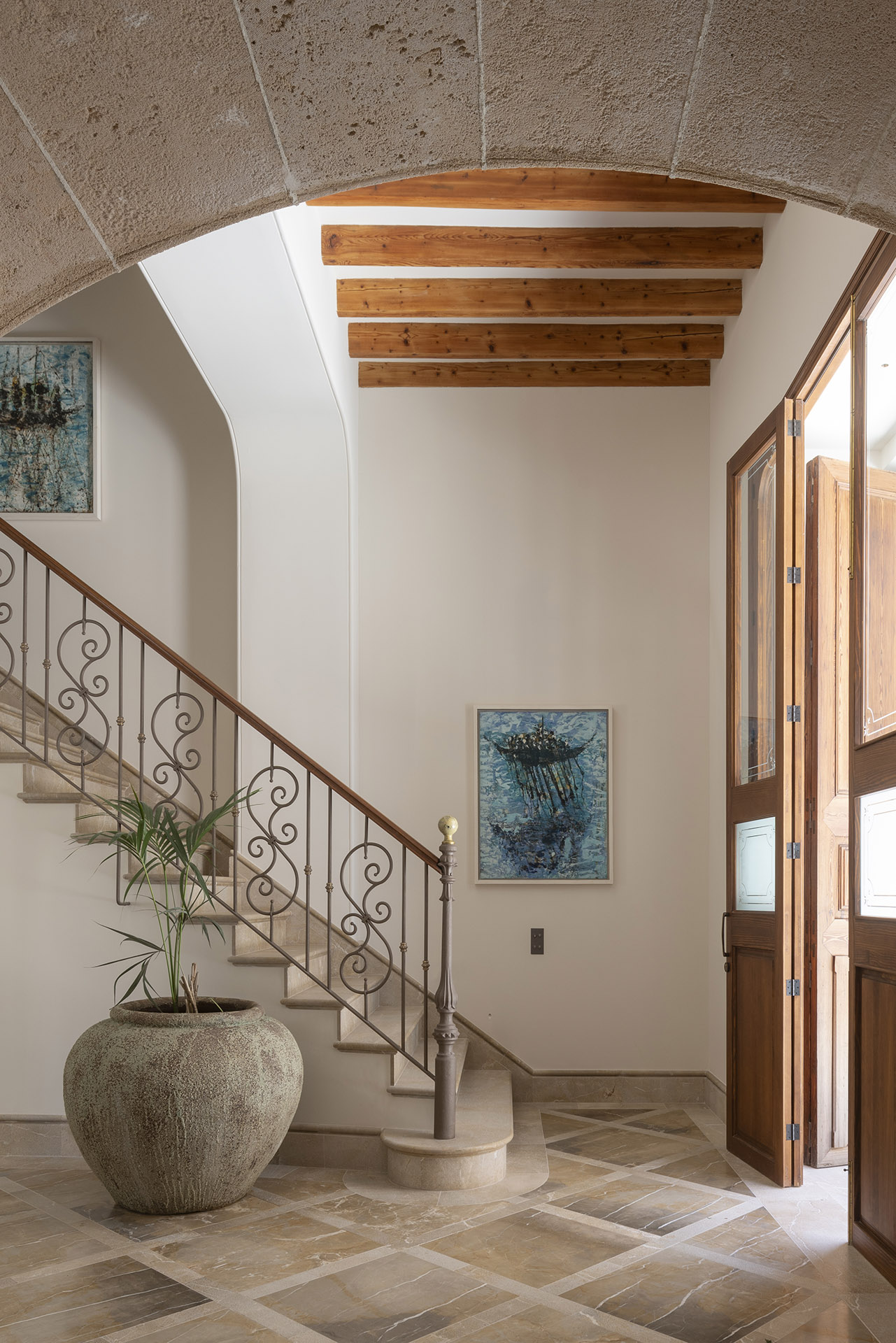 Escalera con barandilla de forja bajo un arco de piedra, cuadros azules y vigas de madera