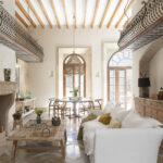 Salón comedor con chimenea de piedra, sofá blanco, vigas de madera y balcones interiores de forja