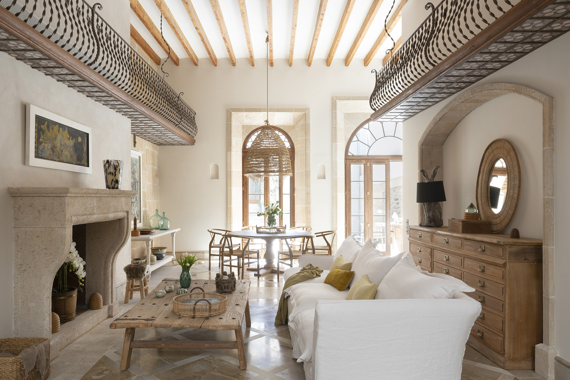 Salón comedor con chimenea de piedra, sofá blanco, vigas de madera y balcones interiores de forja
