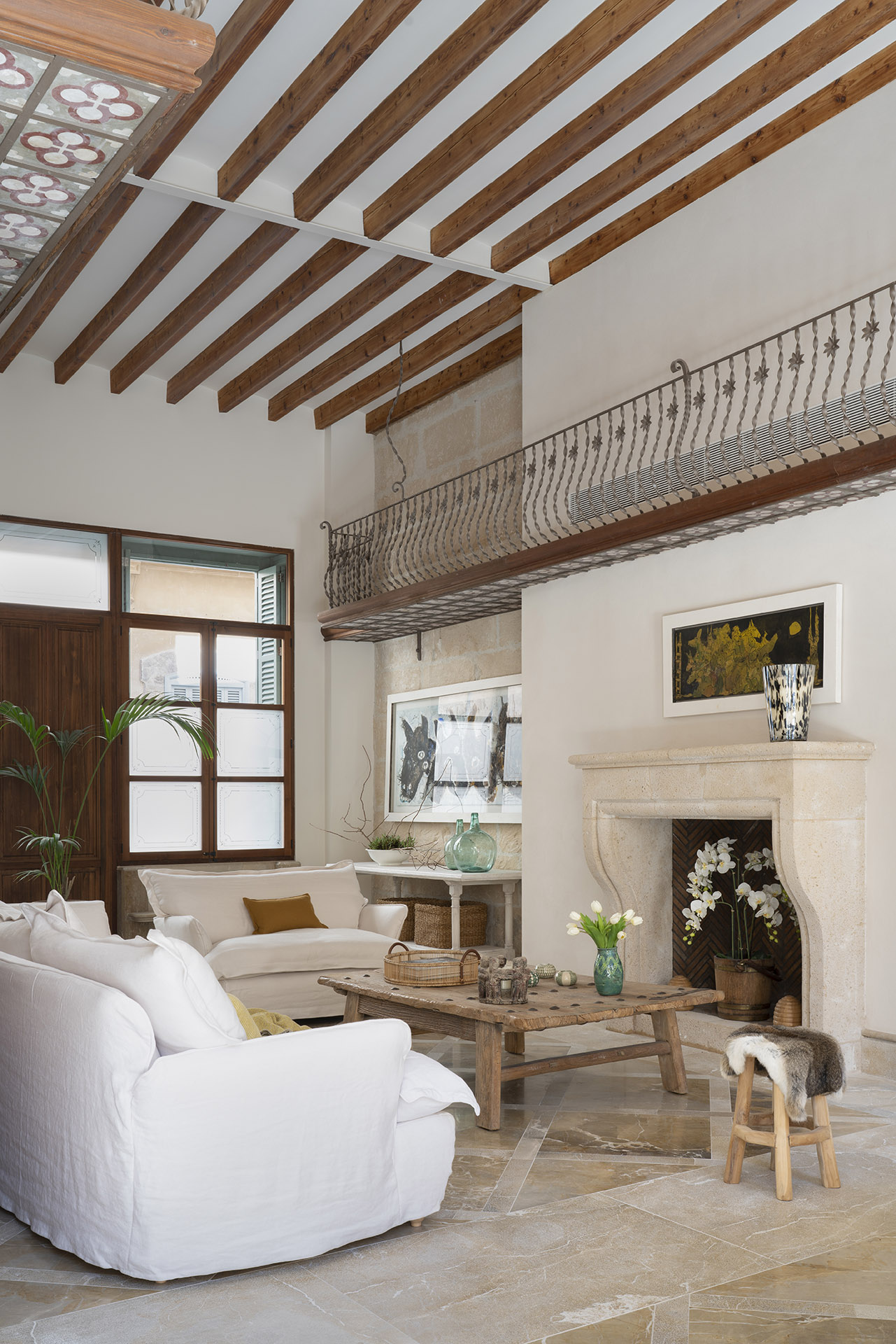 Salón de techos altos con vigas vistas, balcón de forja, chimenea de piedra y sofá blanco
