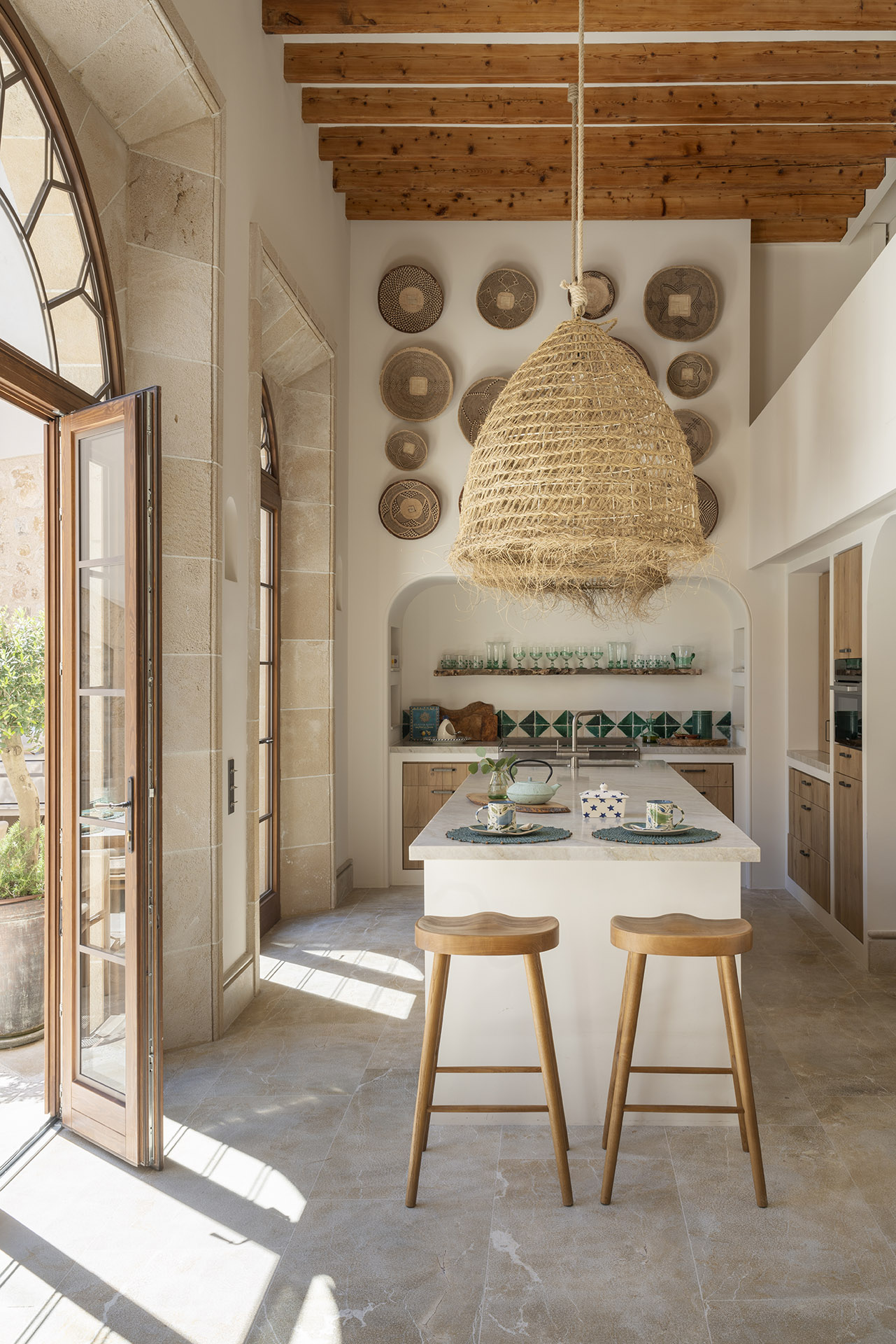 Cocina de techos altos con isla blanca, taburetes de madera, lámpara de esparto y luz natural