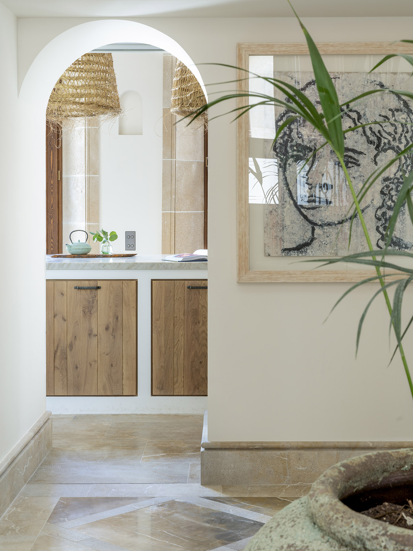 Pasillo con arco blanco que enmarca la cocina, armarios de madera, cuadro con rostro y palmera