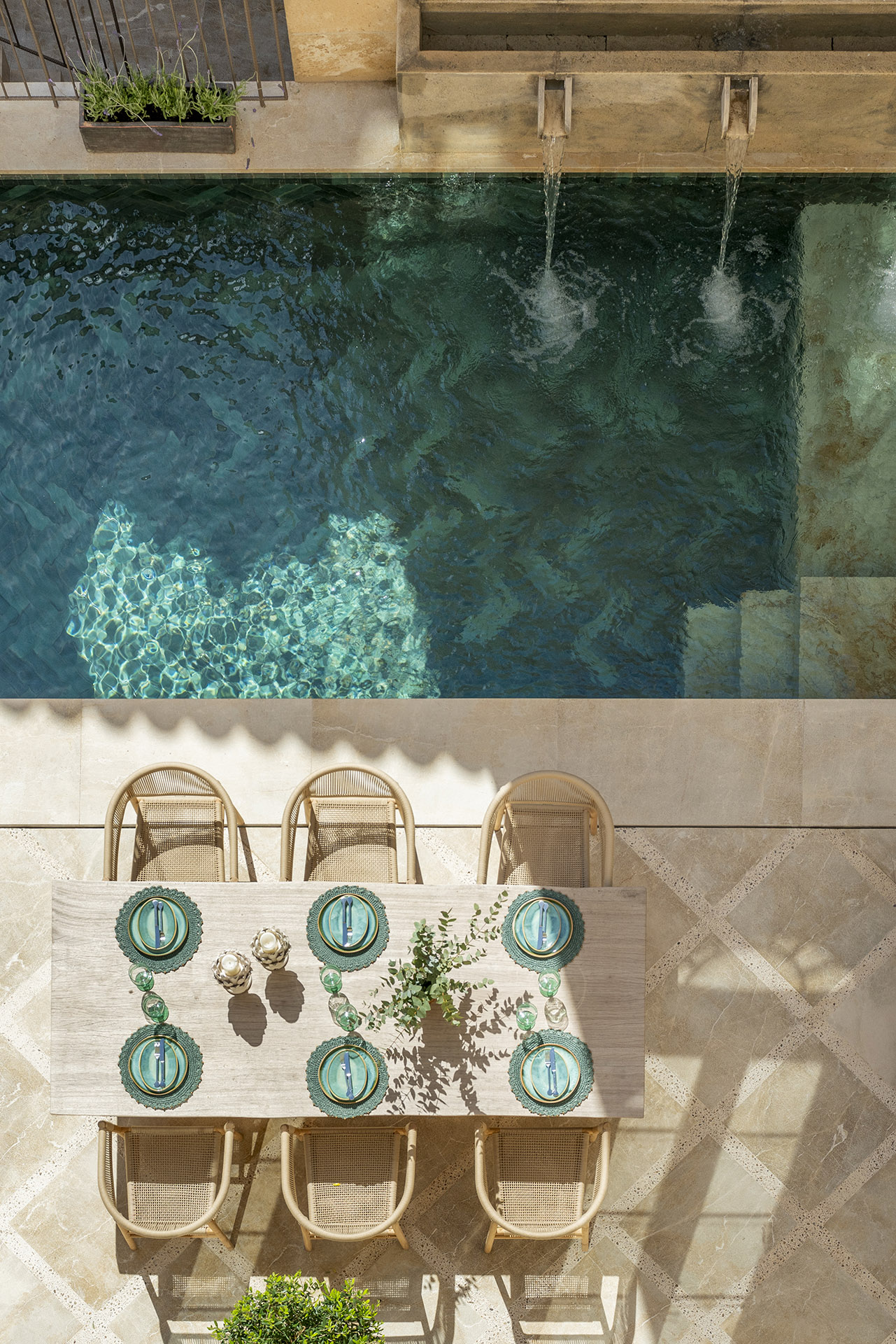 Vista cenital de la piscina con surtidores y mesa puesta con seis sillas sobre suelo de piedra