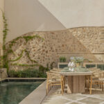 Patio con surtidores sobre la piscina, muro de piedra con enredadera y mesa de comedor exterior