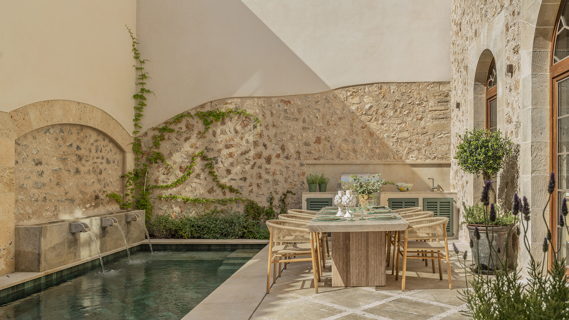 Patio con surtidores sobre la piscina, muro de piedra con enredadera y mesa de comedor exterior