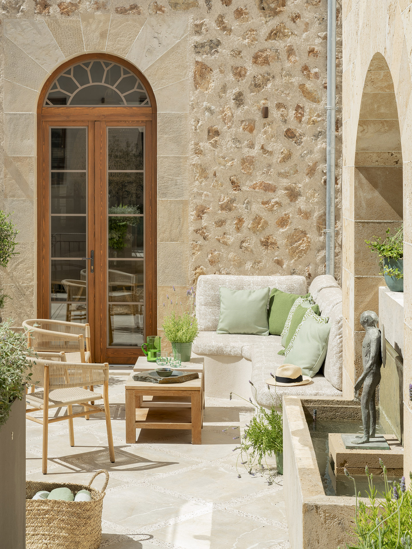 Rincón del patio con banco corrido, cojines verdes, sillas de mimbre, puerta arqueada y escultura