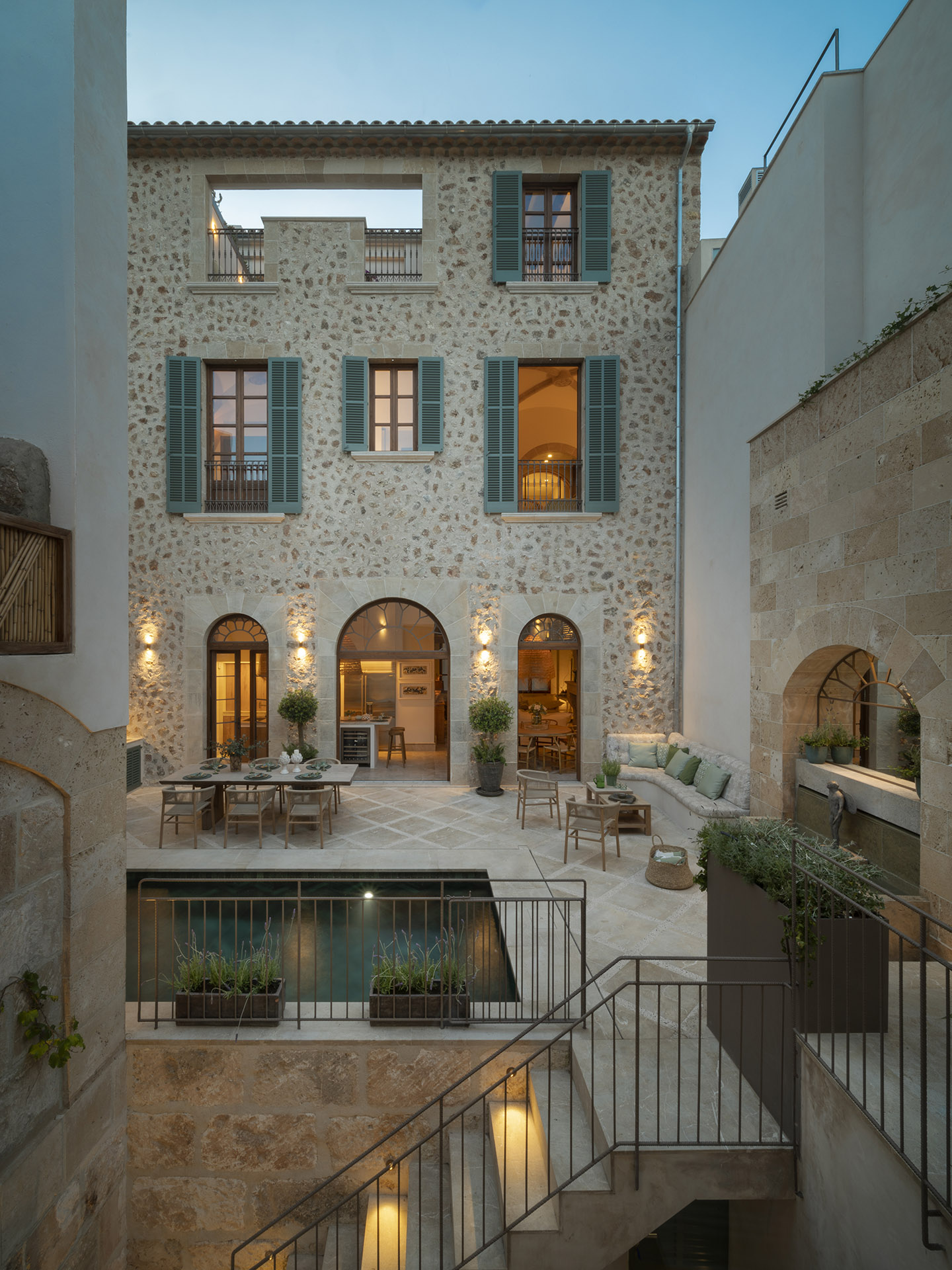 Fachada del patio al anochecer con luz cálida, muros de piedra, contraventanas verdes y piscina