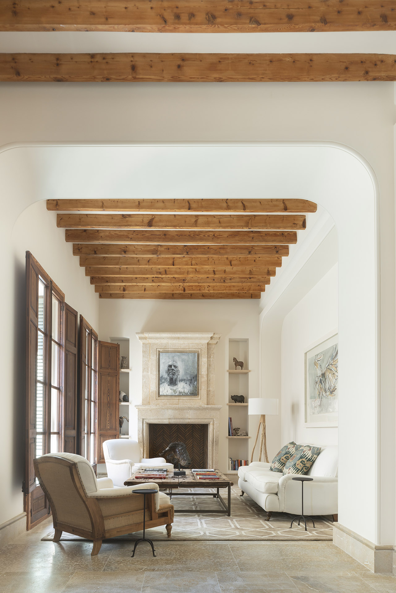 Salón con vigas de madera, arco blanco, chimenea de piedra, butacas y sofá claro