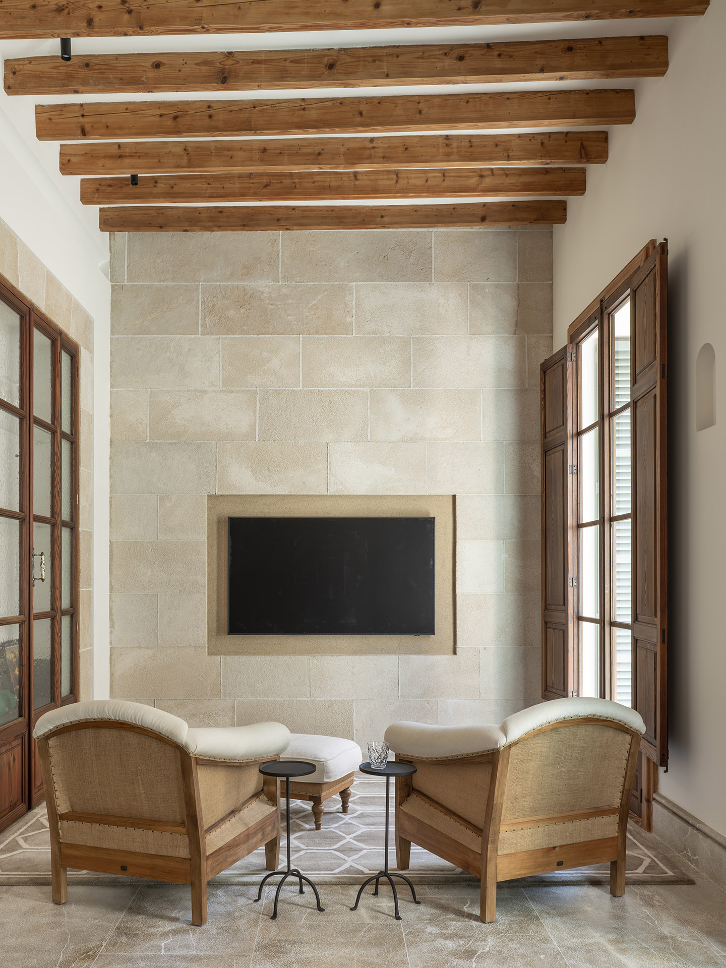 Rincón de estar con pared de piedra, televisor empotrado, dos butacas tapizadas y vigas de madera