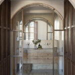 Baño tras una puerta acristalada en arco, con lavabo doble de madera y pavimento de piedra