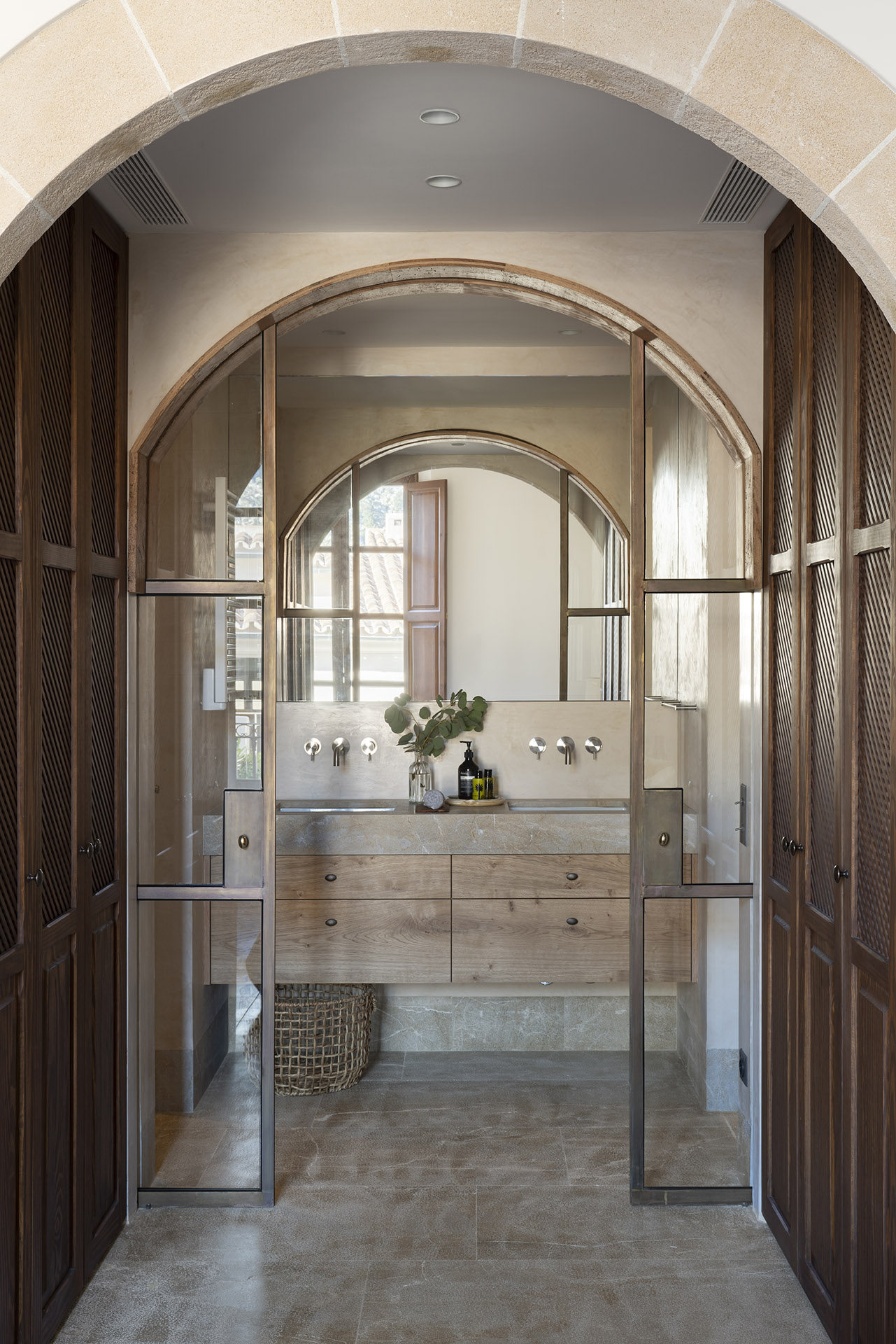 Baño tras una puerta acristalada en arco, con lavabo doble de madera y pavimento de piedra