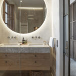 Baño con espejo redondo retroiluminado, lavabo doble con encimera de piedra y mueble de madera