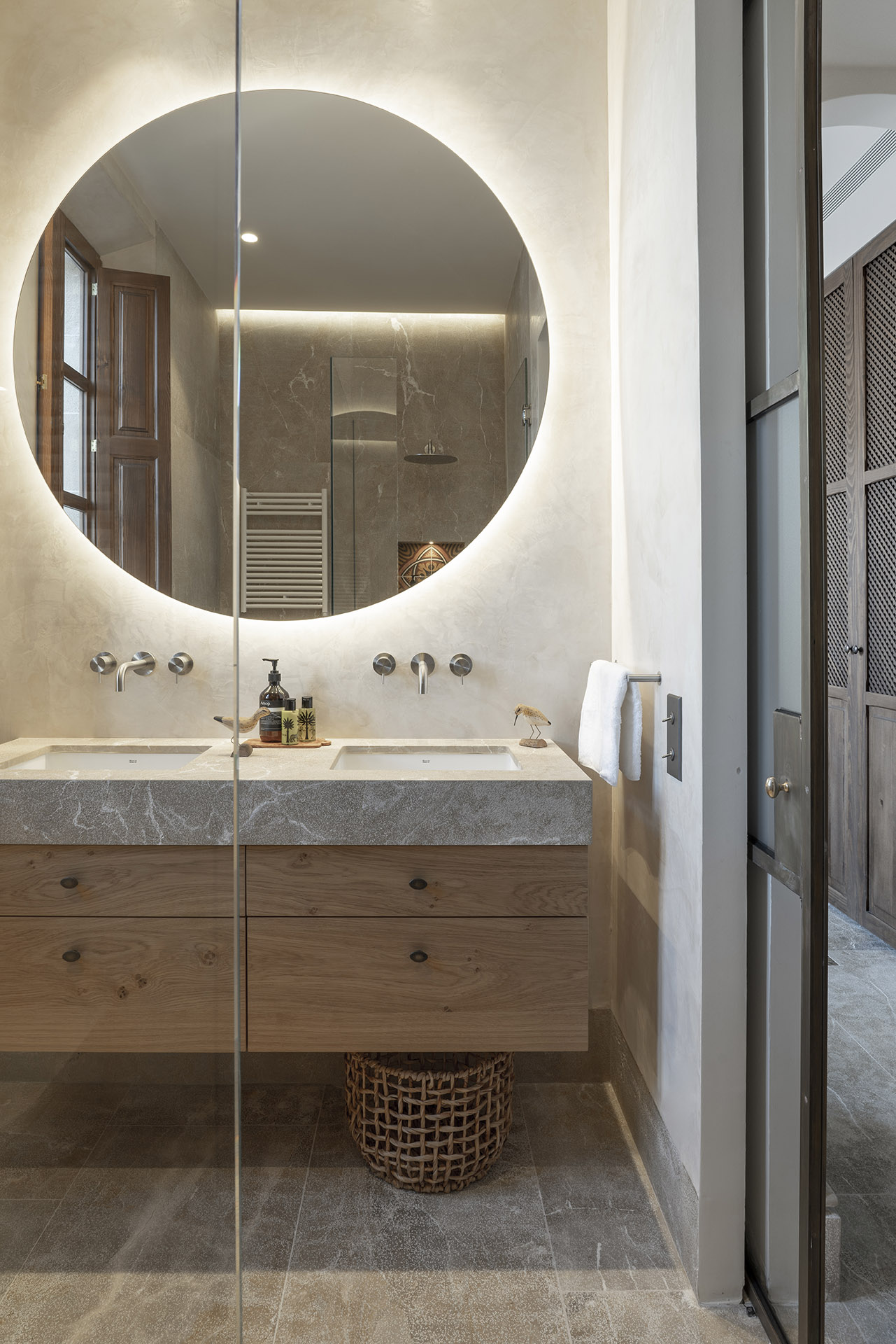 Baño con espejo redondo retroiluminado, lavabo doble con encimera de piedra y mueble de madera