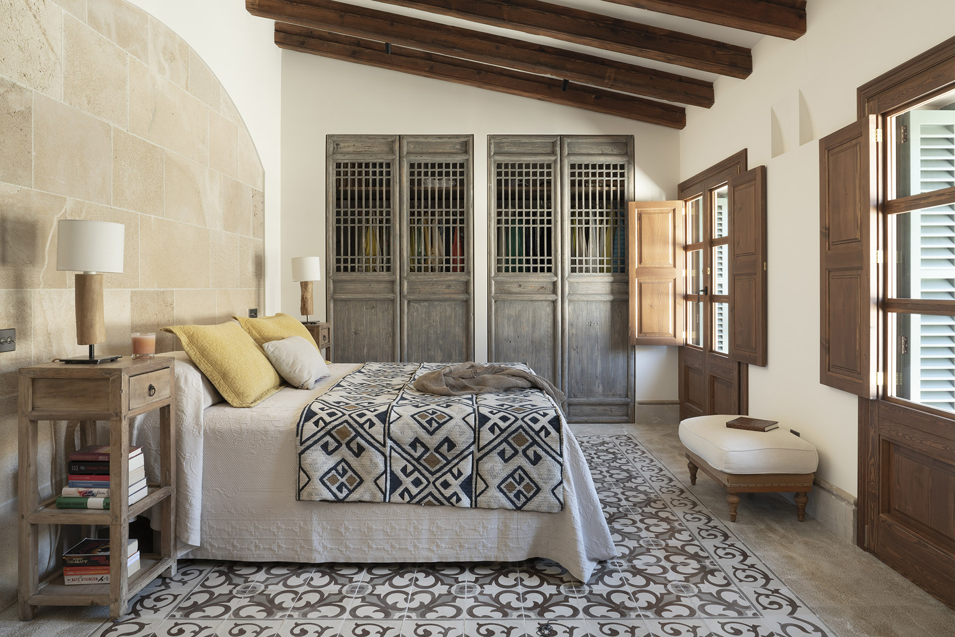 Dormitorio con pared de piedra, armarios de madera con celosía, colcha geométrica y suelo de baldosa decorativa