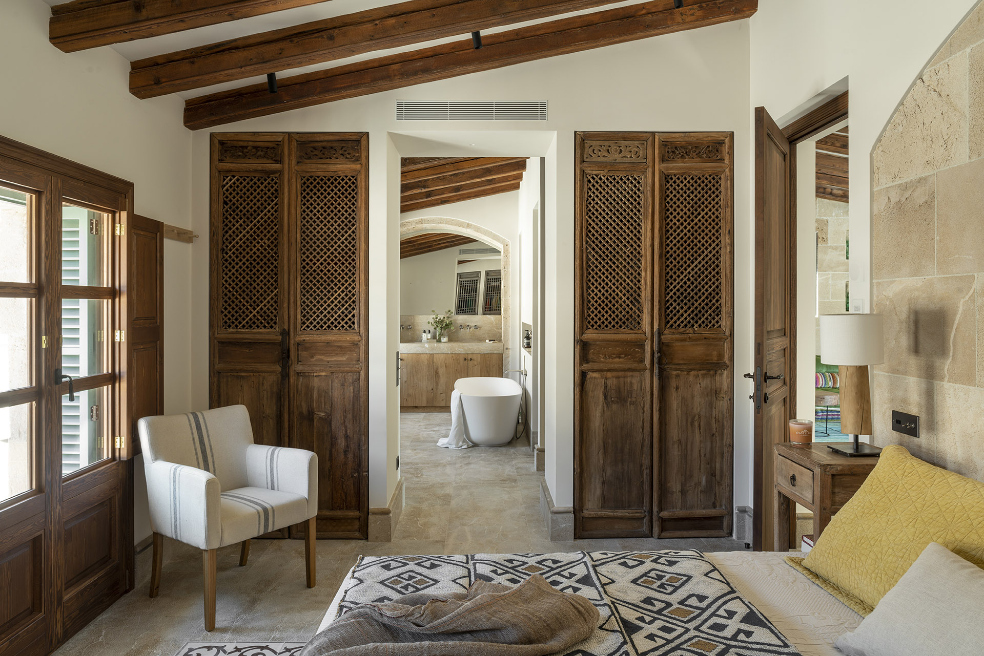 Dormitorio con puertas de celosía de madera y, al fondo, baño con bañera exenta bajo vigas