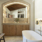 Baño con bañera exenta blanca, lavabo doble de madera y espejo enmarcado por arco de piedra iluminado