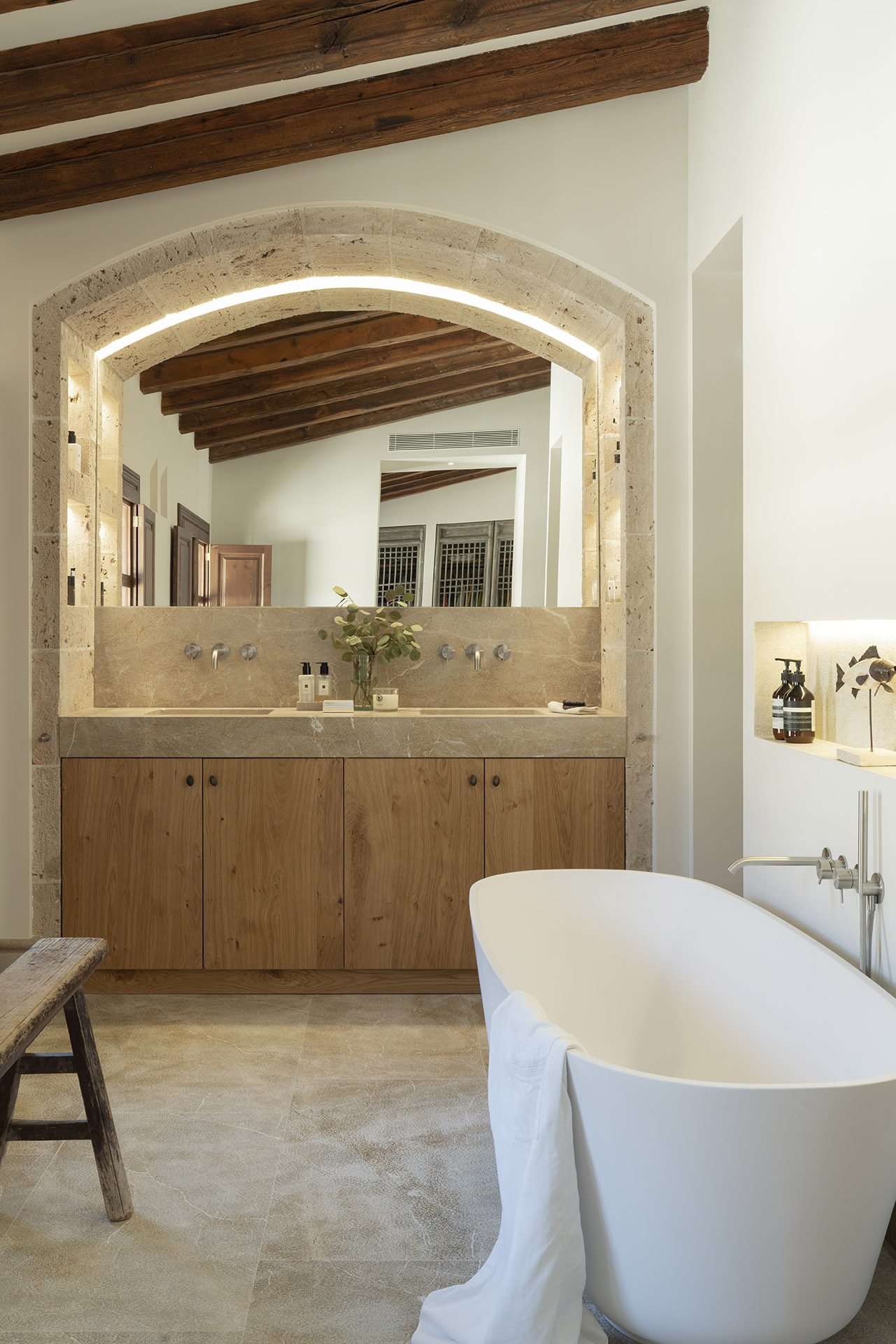 Baño con bañera exenta blanca, lavabo doble de madera y espejo enmarcado por arco de piedra iluminado
