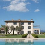 Fassade des Herrenhauses mit grauen Fensterläden, Olivenbäumen, Pool im Vordergrund und Meer im Hintergrund