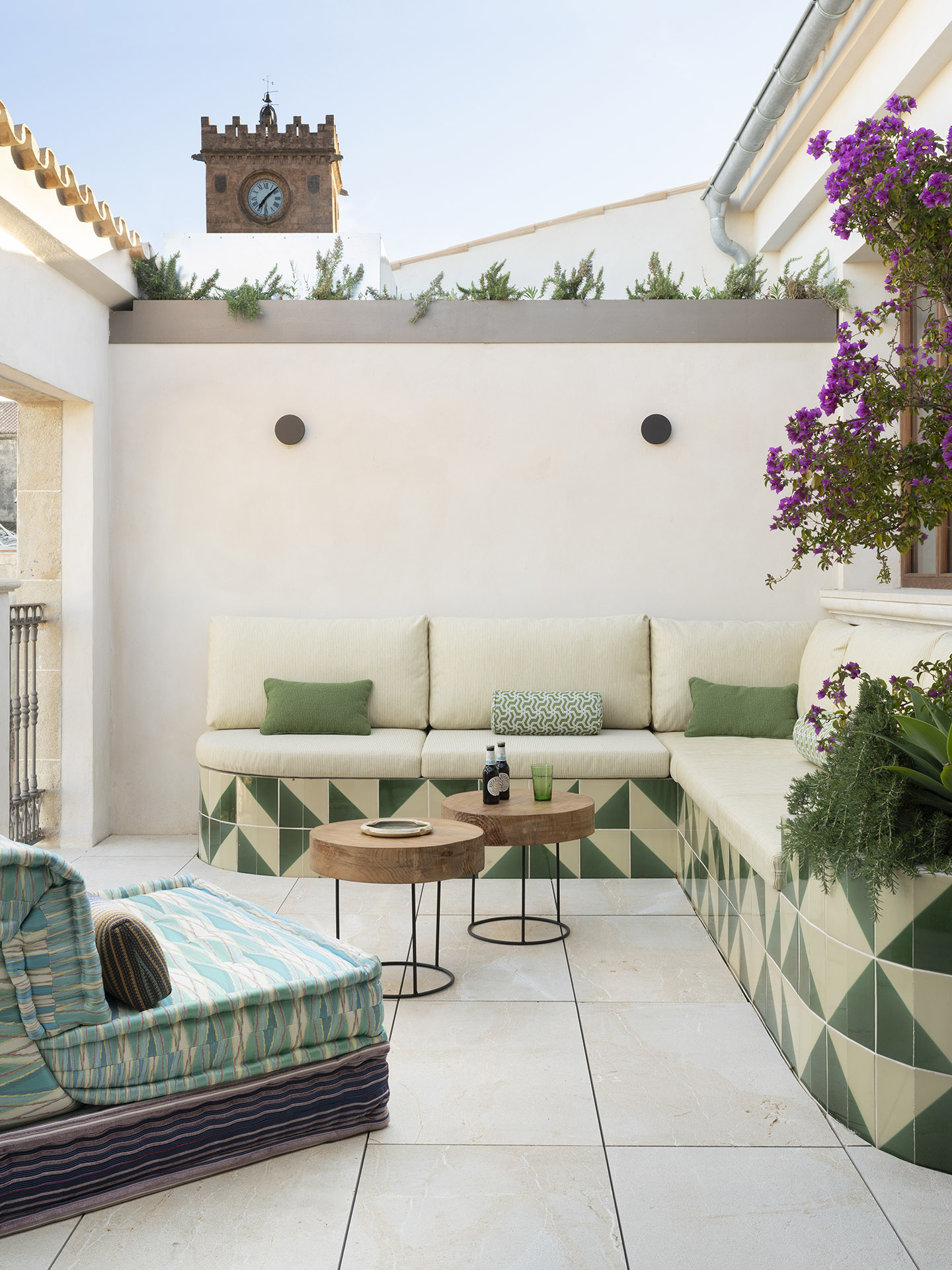 Terraza en altura con banco revestido de azulejos verdes, mesas de madera, buganvilla y torre con reloj