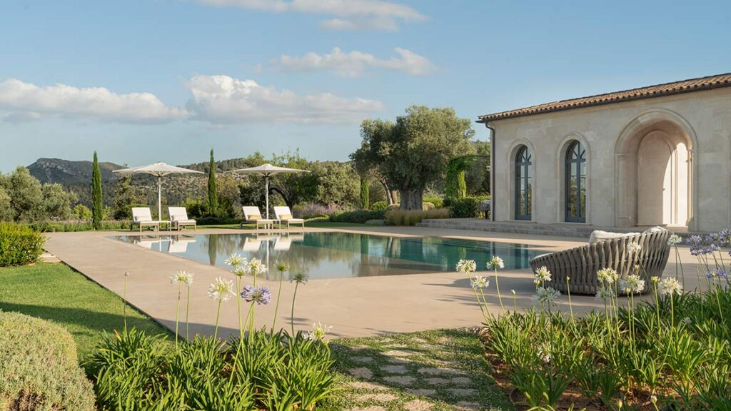 Pool mit Sonnenliegen und Sonnenschirmen neben dem Steinpavillon, blühende Schmucklilien und Berge im Hintergrund