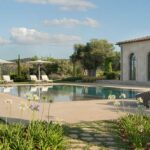 Pool mit Sonnenliegen und Sonnenschirmen neben dem Steinpavillon, blühende Schmucklilien und Berge im Hintergrund