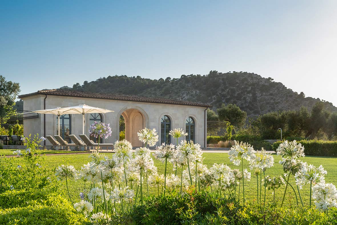 Weiß blühende Schmucklilien im Vordergrund und Steinpavillon mit Sonnenliegen vor einem bewaldeten Hügel