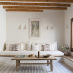 Salón con banco corrido blanco, mesa baja de madera, vigas vistas y gran ventana de madera