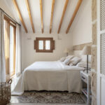 Dormitorio con vigas de madera, cama blanca, suelo de baldosa hidráulica y pared de piedra vista