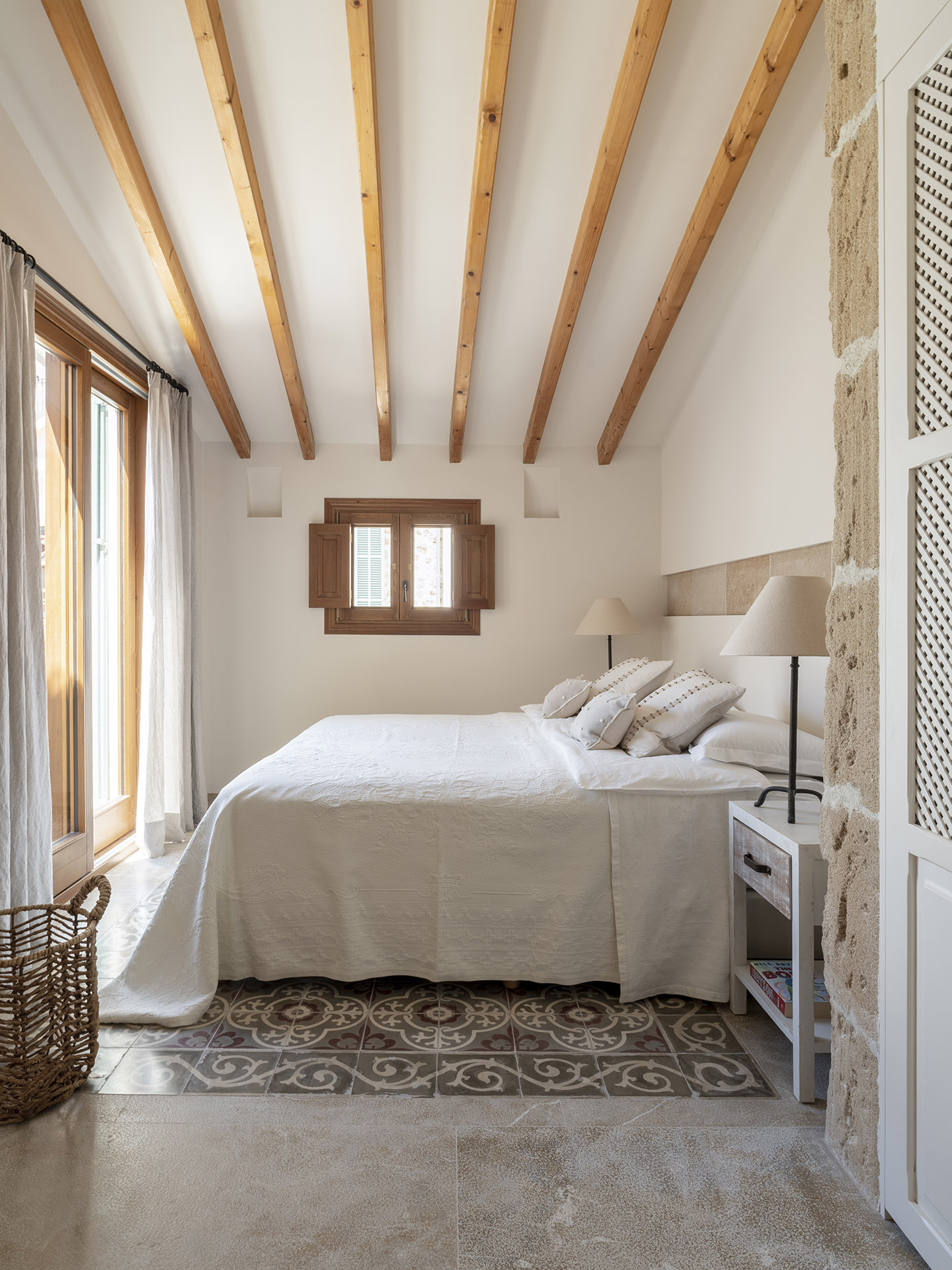 Dormitorio con vigas de madera, cama blanca, suelo de baldosa hidráulica y pared de piedra vista