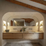 Baño con lavabo doble sobre mueble de madera, espejo en arco, hornacinas iluminadas y suelo de piedra