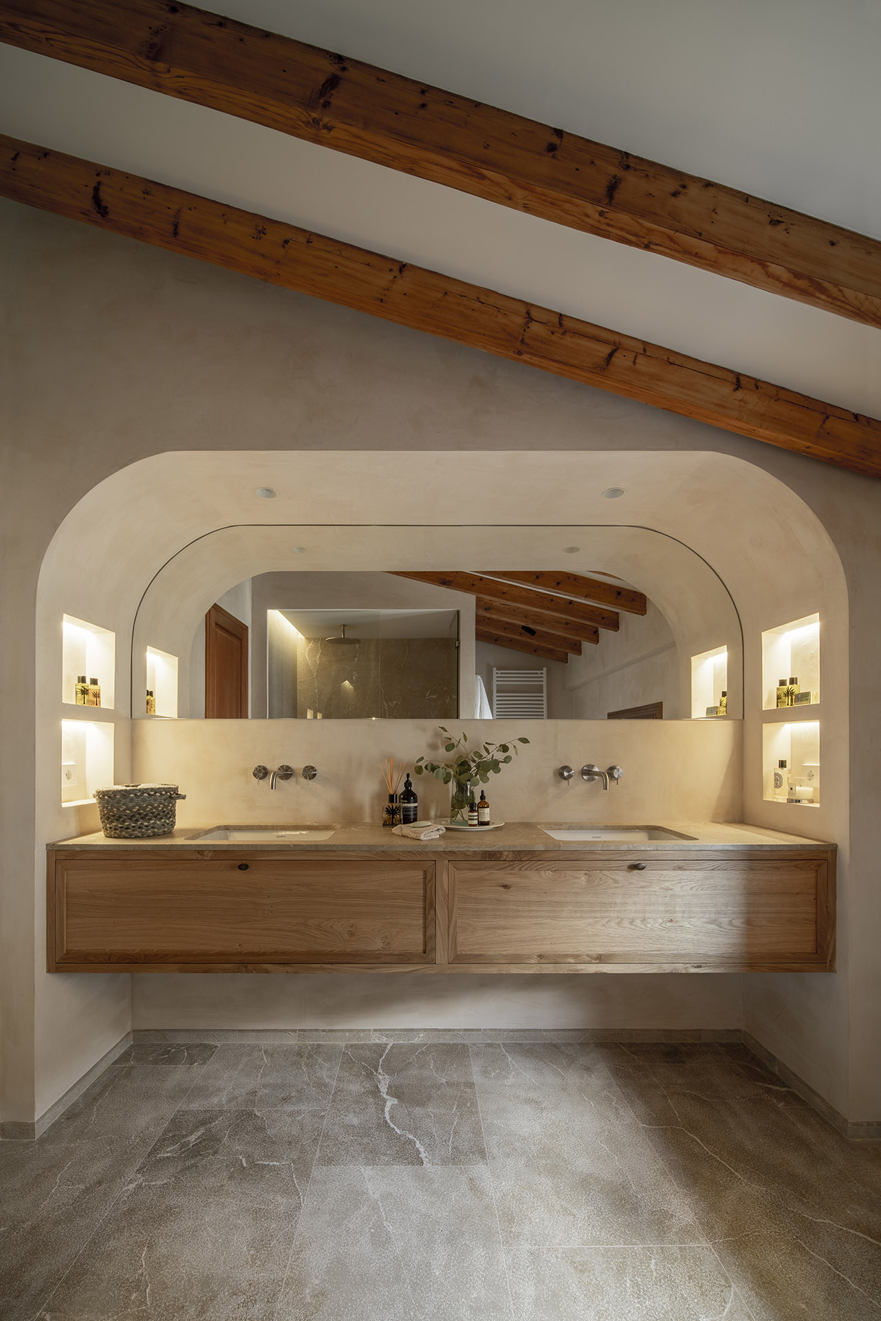 Baño con lavabo doble sobre mueble de madera, espejo en arco, hornacinas iluminadas y suelo de piedra