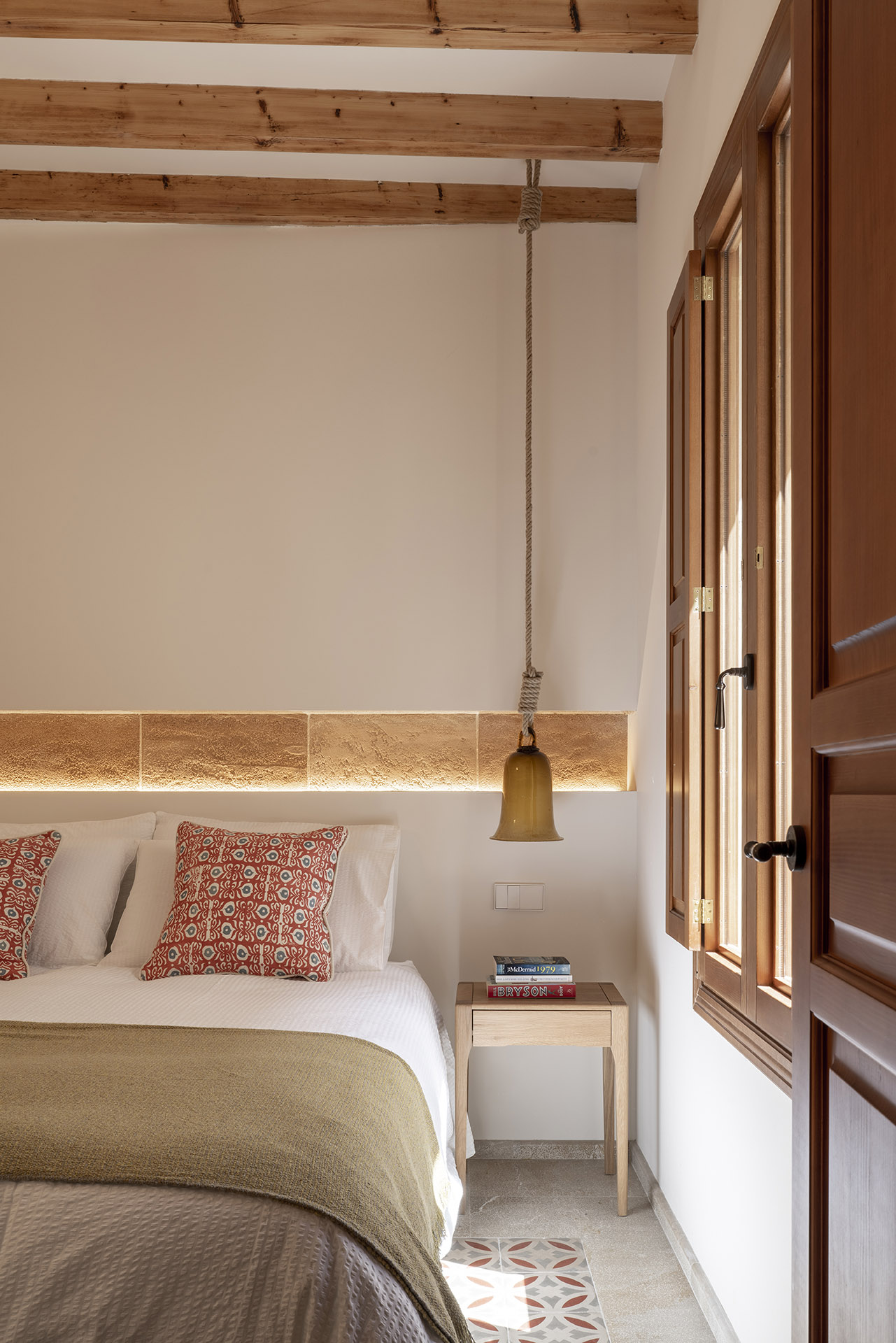 Dormitorio con franja de piedra iluminada, lámpara colgante de campana, cojines rojos y ventana de madera