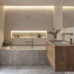 Baño con bañera empotrada revestida de piedra, hornacina iluminada, mueble de madera y paredes lisas color arena