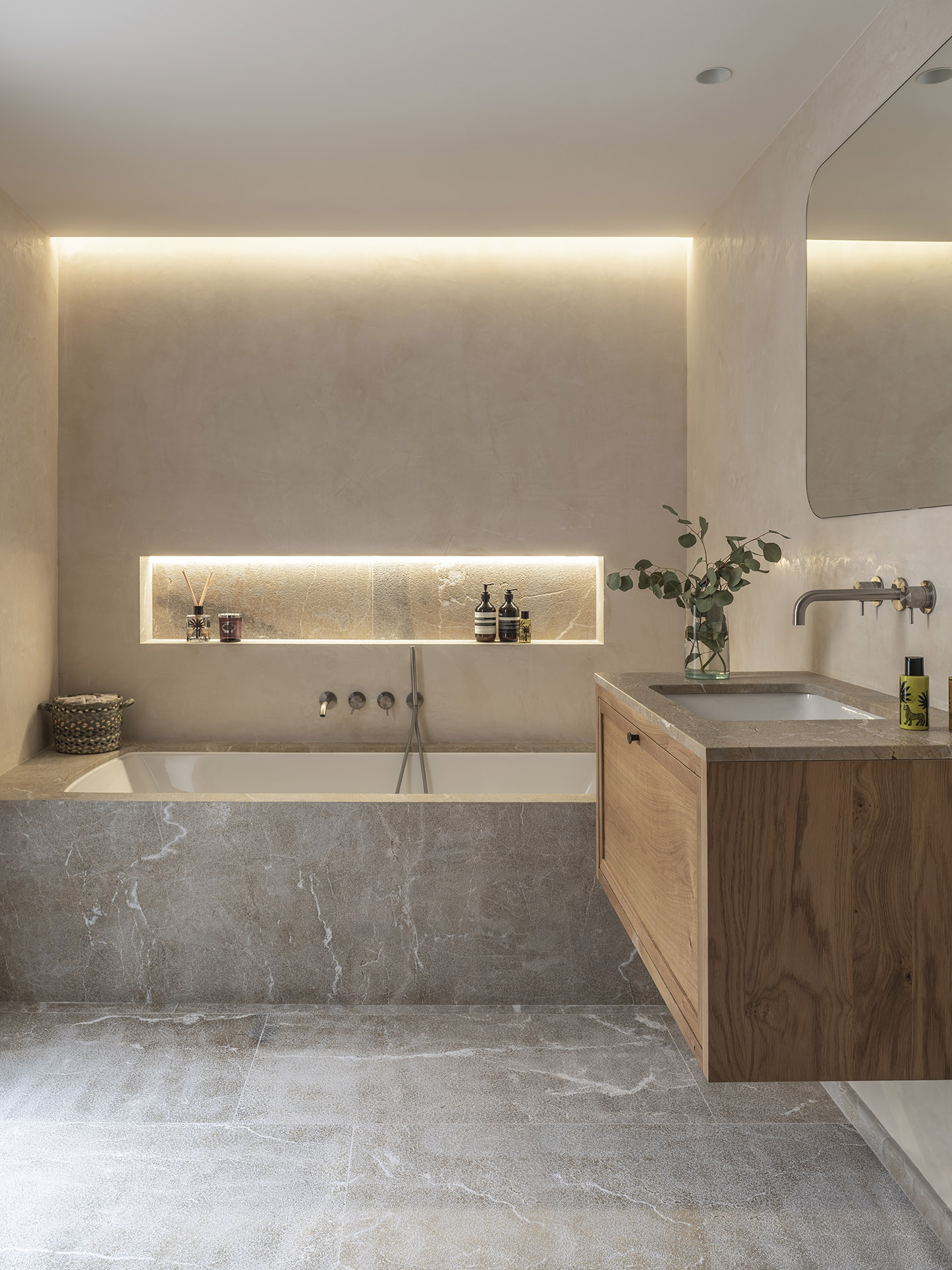 Baño con bañera empotrada revestida de piedra, hornacina iluminada, mueble de madera y paredes lisas color arena