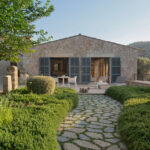 Casita de piedra con contraventanas grises abiertas, sendero empedrado y setos redondeados en el jardín