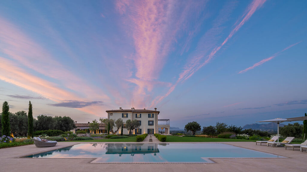 Poolbereich mit Sonnenliegen und Sonnenschirmen, dahinter das Haus unter rosa Himmel in der Dämmerung