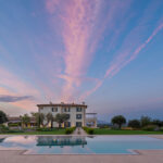 Poolbereich mit Sonnenliegen und Sonnenschirmen, dahinter das Haus unter rosa Himmel in der Dämmerung